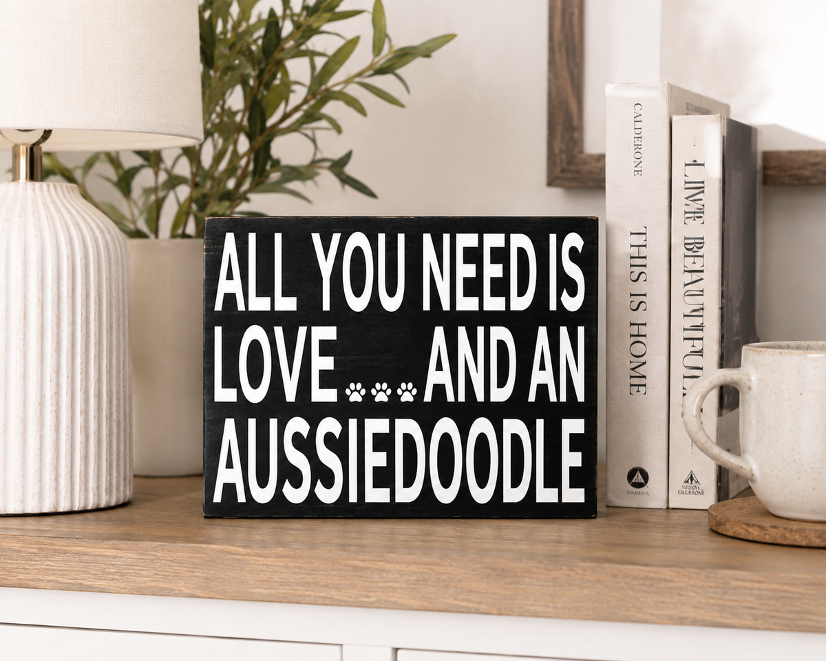 A rectangular black sign with white text reading 'ALL YOU NEED IS LOVE *** AND AN AUSSIEDOOLE' displayed on a wooden shelf next to books, a lamp, and a mug.
