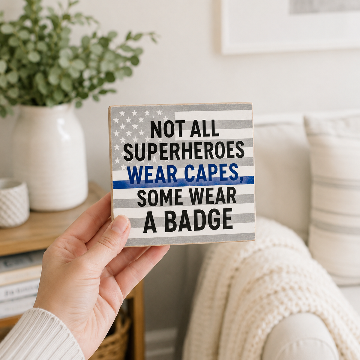 A hand holds a square wooden sign with a distressed American flag design and the text 'NOT ALL SUPERHEROES WEAR CAPES SOME WEAR A BADGE' in black and blue lettering. The background shows a cozy living room setting with a sofa, throw blanket, and potted plant.
