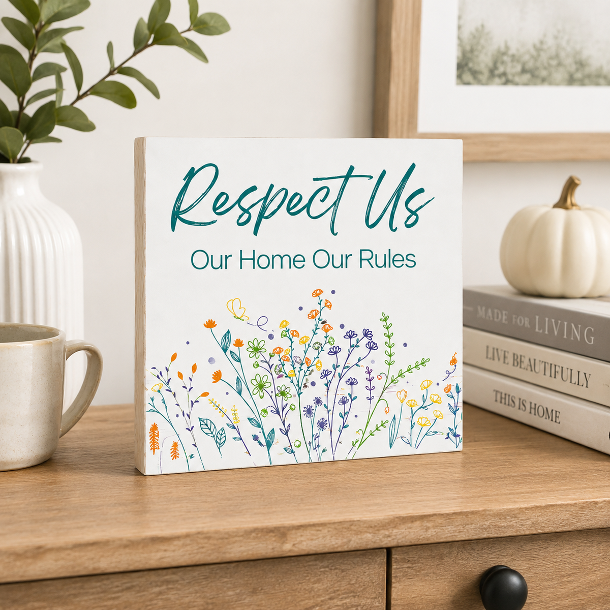 A square decorative sign with the phrase 'Respect Us Our Home Our Rules' in teal lettering above a colorful wildflower illustration, displayed on a wooden table in a styled home setting with a mug, books, and a small pumpkin.