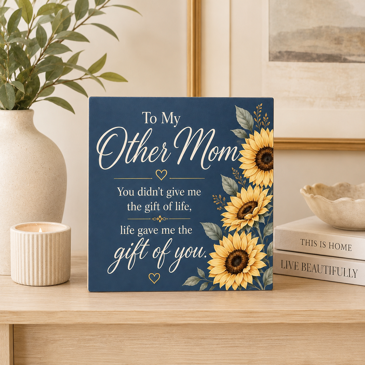A square decorative sign with a navy blue background and yellow sunflower illustrations, placed on a light wood table in a styled home setting. The sign reads, 'To My Other Mom You didn't give me the gift of life, life gave me the gift of you.