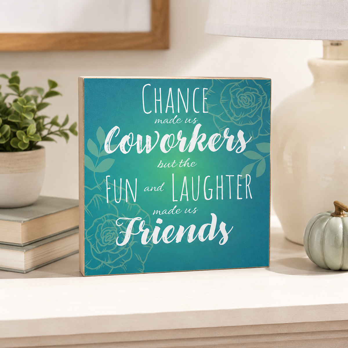 A square decorative sign with a teal and blue floral background and white text reading, 'Chance made us coworkers but the fun and laughter made us friends,' styled on a tabletop with a plant, books, a lamp, and a small pumpkin.