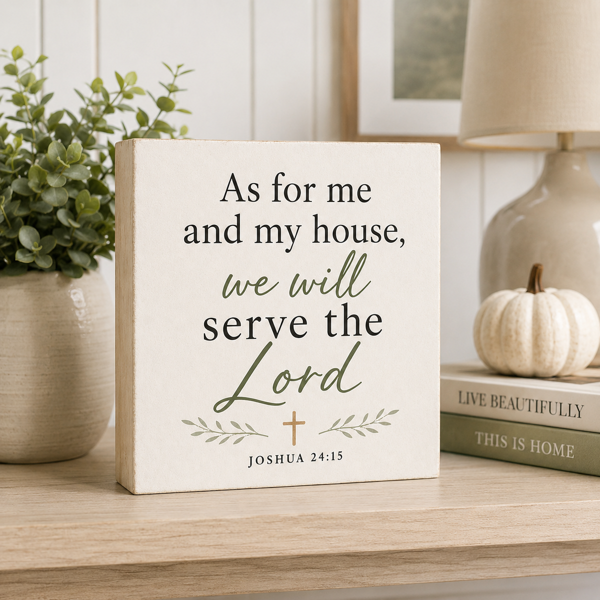 A square cream-colored wooden sign with the Bible verse 'As for me and my house, we will serve the Lord Joshua 24:15' in dark green and black lettering, styled on a light wood shelf next to a potted plant and a small white pumpkin.