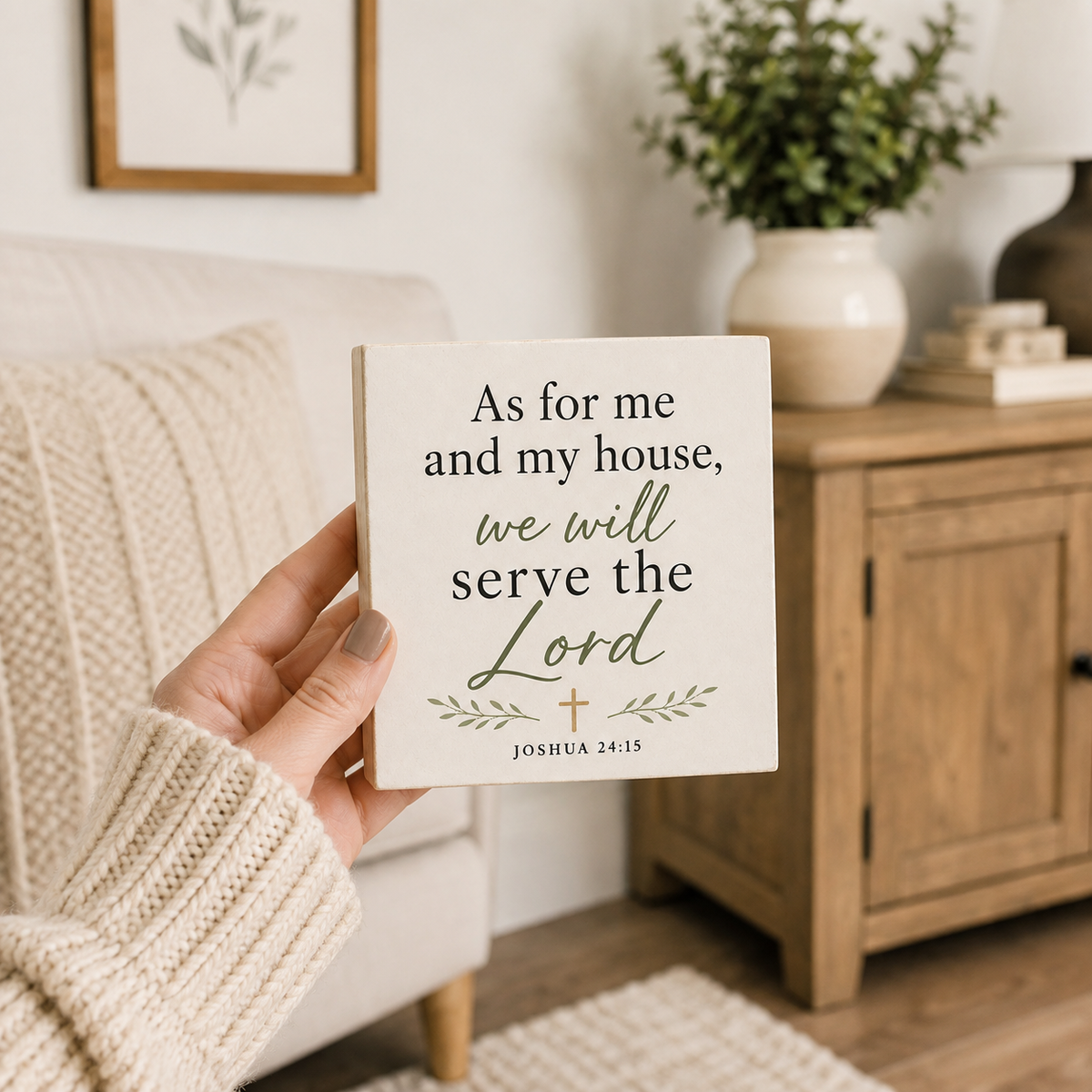 A hand in a cream knit sweater holds a square white decorative sign with the Bible verse 'As for me and my house, we will serve the Lord' in black and green lettering. The background shows a cozy, softly blurred living room setting with a wooden cabinet, potted plant, and framed art.