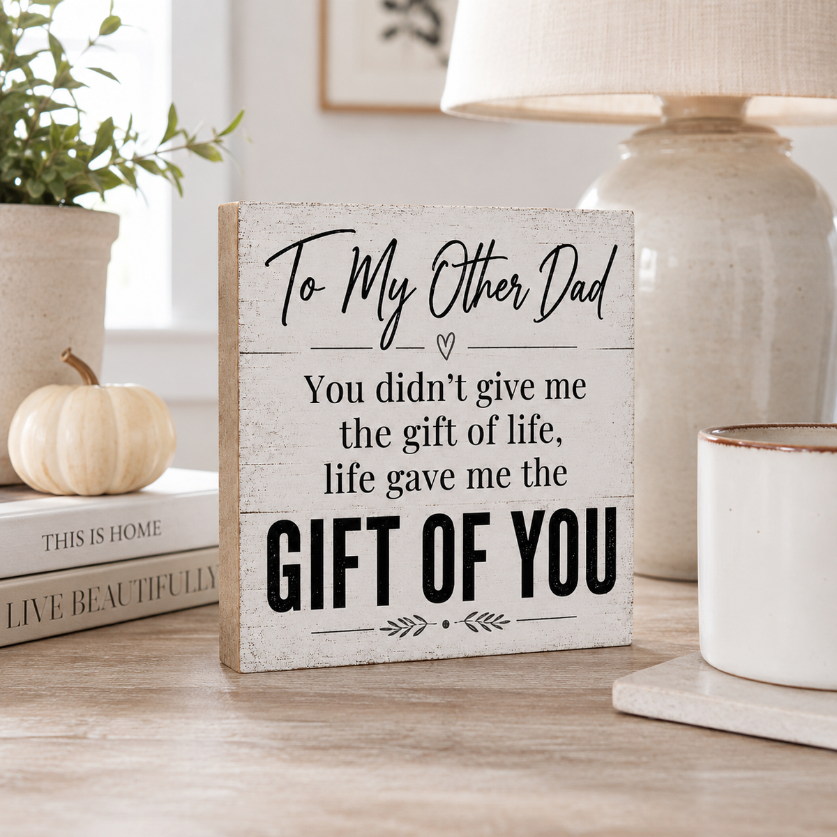 A rustic white wooden sign with black text reading 'To My Other Dad You didn't give me the gift of life, life gave me the GIFT OF YOU' stands on a wooden table in a styled home setting with a lamp, mug, books, and a small pumpkin.