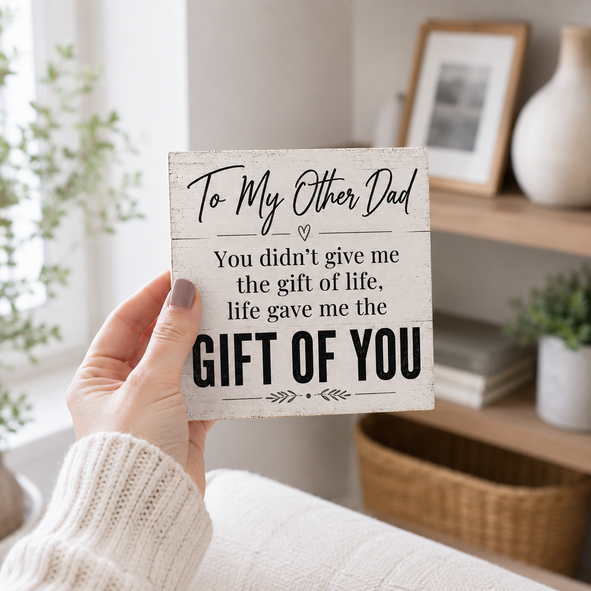 A hand wearing a cream knit sweater holds a rustic white wooden sign with black text that reads, 'To My Other Dad You didn't give me the gift of life, life gave me the GIFT OF YOU.' The background shows a softly blurred home interior with a shelf, framed picture, and plants.