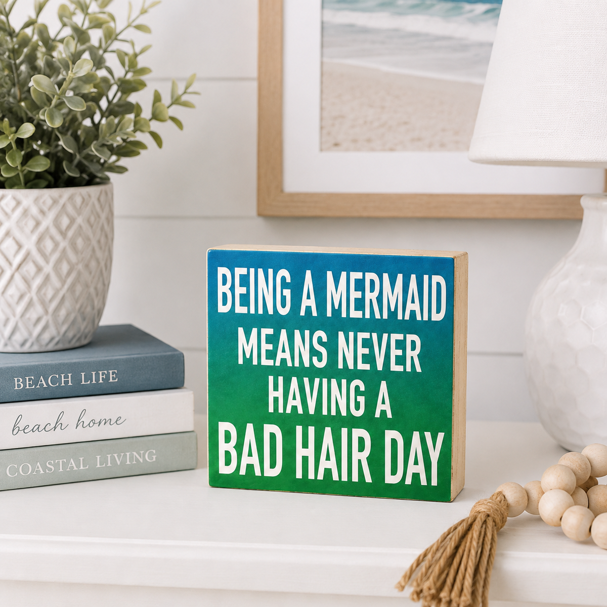 A square wooden sign with the phrase 'BEING A MERMAID MEANS NEVER HAVING A BAD HAIR DAY' in white text over a green-to-teal gradient, styled on a white shelf with a potted plant, books, a lamp, and a bead tassel.