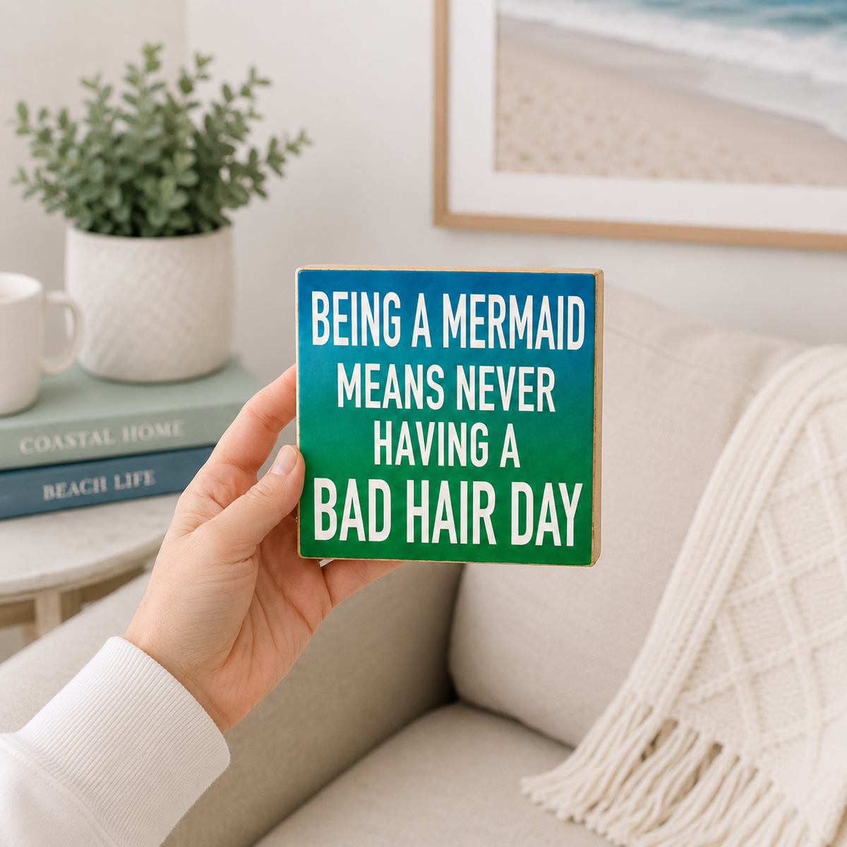 A hand holds a square wooden sign with a teal-to-green gradient background and white text that reads, 'BEING A MERMAID MEANS NEVER HAVING A BAD HAIR DAY.' The scene is a cozy living room with a beige sofa, a plant, books, and framed wall art in the background.