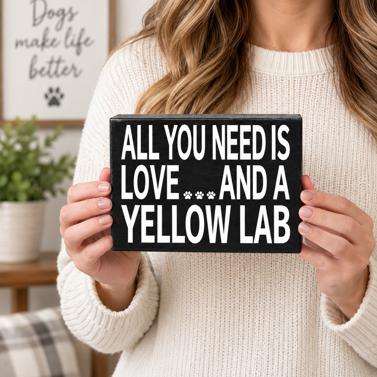 A woman in a cream knit sweater holds a black rectangular sign with bold white text that reads, 'ALL YOU NEED IS LOVE.