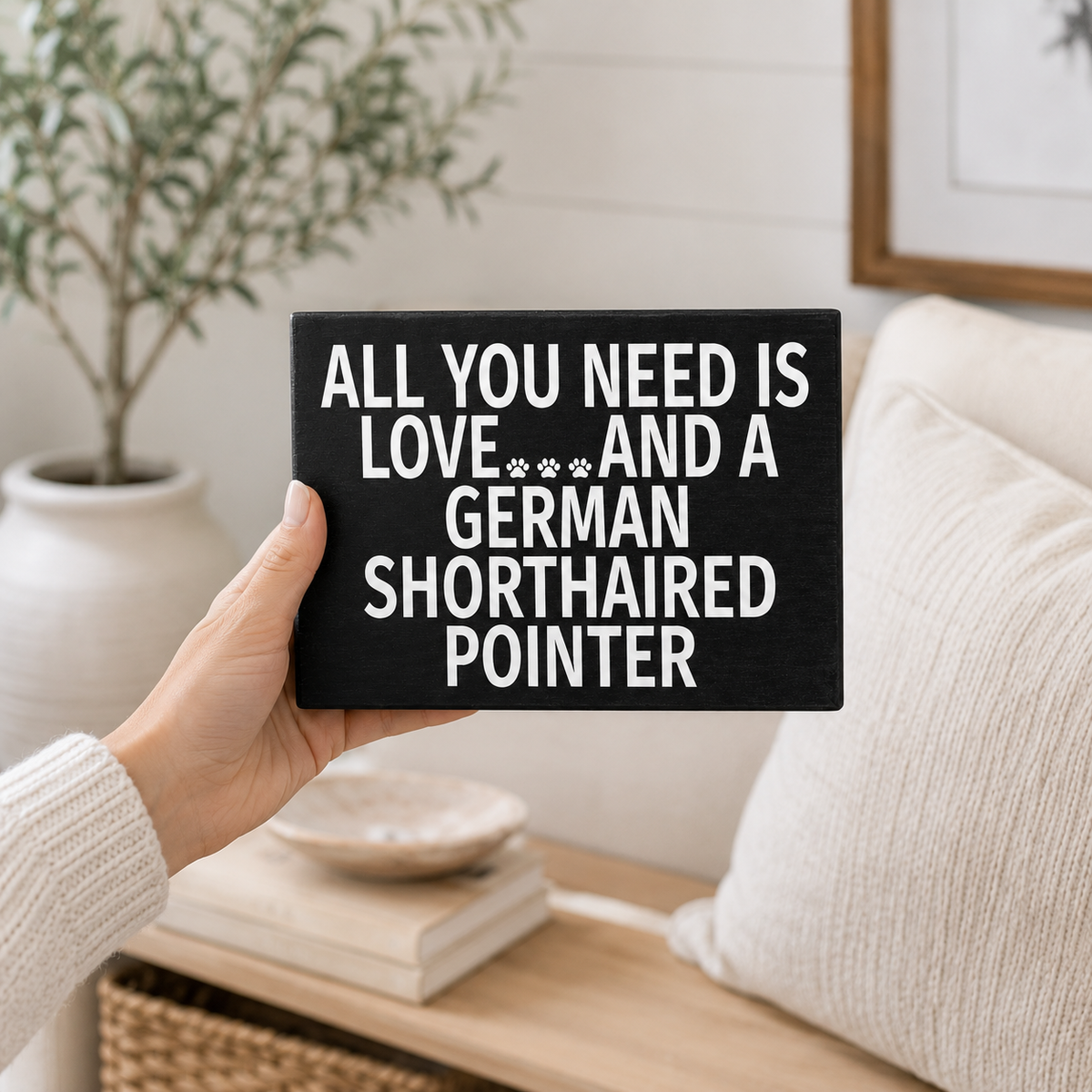 A hand holding a black rectangular sign with white text that reads, 'ALL YOU NEED IS LOVE***AND A GERMAN SHORTHAIRED POINTER' in a cozy, modern living room setting.