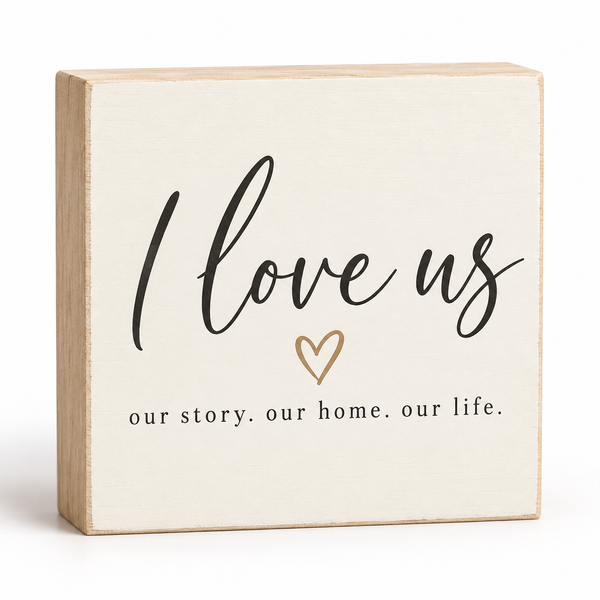 A square decorative wooden block sign with a light natural wood frame and a cream face. The face features the phrase 'I love us' in black script, a small gold heart outline, and the words 'our story.
