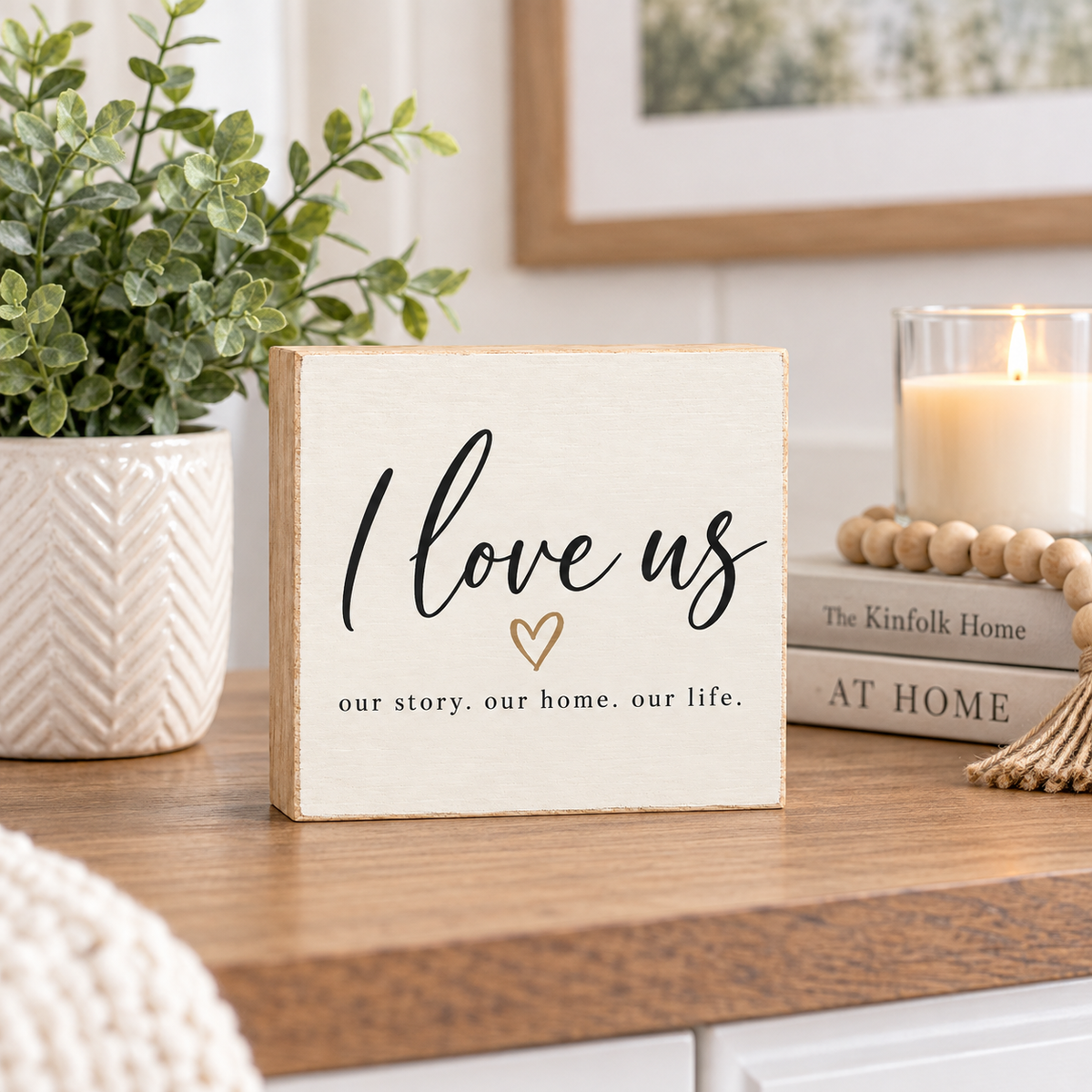 A square cream-colored wooden sign with a rustic finish stands on a wooden table. The sign features the phrase 'I love us' in black script with a small gold heart and the words 'our story.