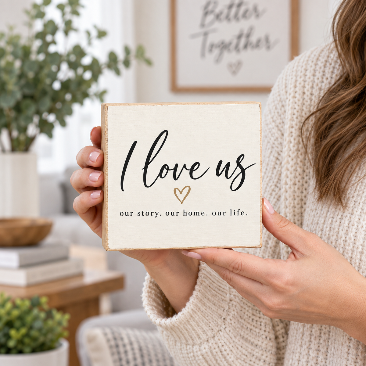 A person in a cream knit sweater holds a square wooden sign that reads 'I love us' with a small gold heart and the phrase 'our story. our home.