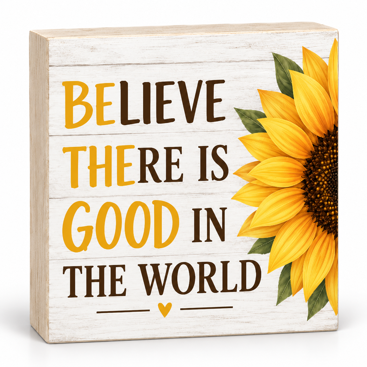 A square decorative wooden sign with a whitewashed wood texture, featuring the text 'BELIEVE THERE IS GOOD IN THE WORLD' in yellow and dark brown lettering, alongside a large yellow sunflower illustration on the right side.
