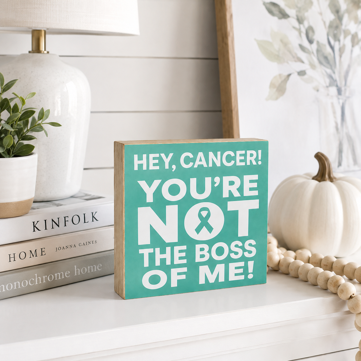 A square wooden sign with a teal front and natural wood sides reads 'HEY, CANCER! YOU'RE NOT THE BOSS OF ME!' in white text with a teal ribbon symbol. It is styled on a white shelf next to a small white pumpkin, wooden beads, a lamp, and books.