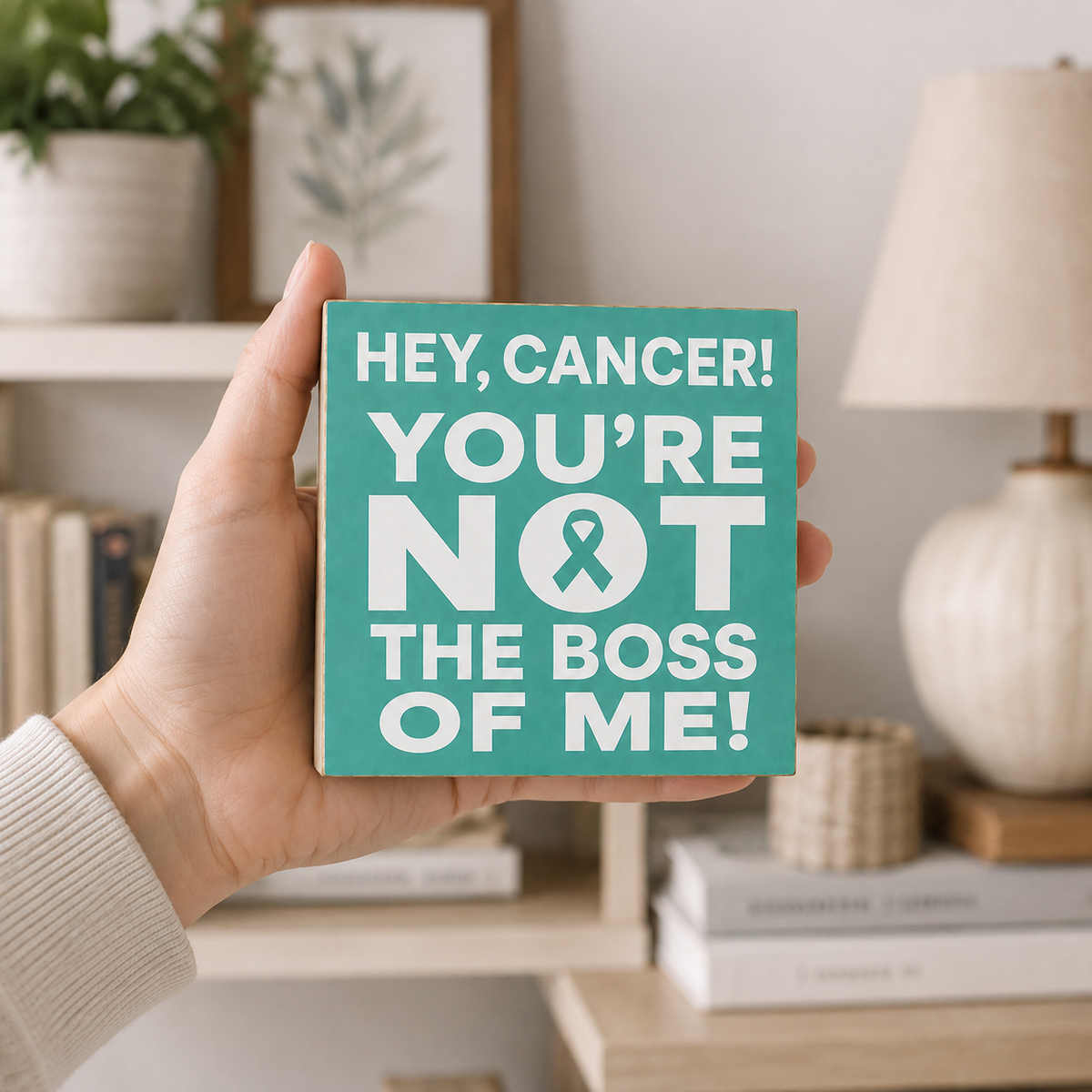 A hand holds a square teal sign with bold white text reading 'HEY, CANCER! YOU'RE NOT THE BOSS OF ME!' with a ribbon symbol, set against a softly blurred home interior background.
