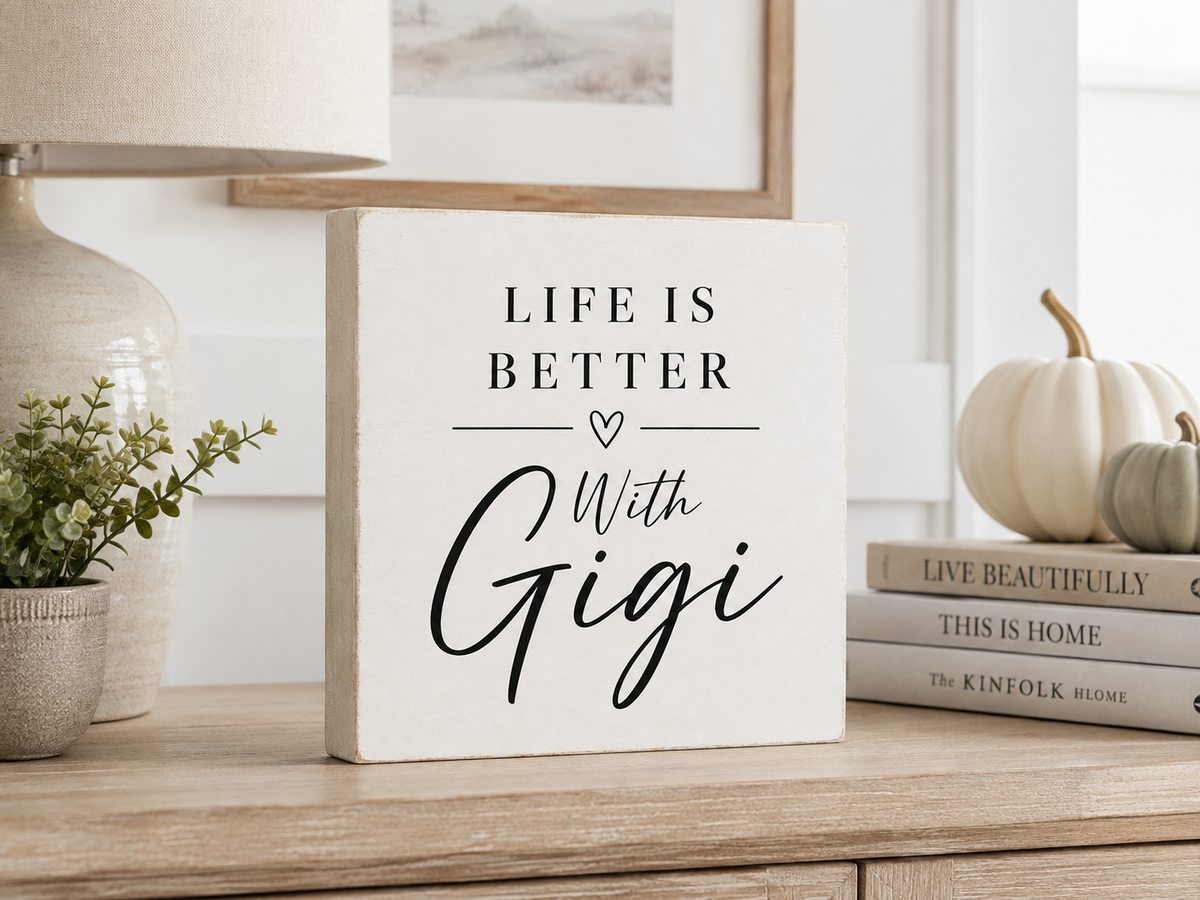 A square white decorative sign with black text reading 'LIFE IS BETTER With Gigi' sits on a wooden table in a styled home interior. A lamp, small plant, books, and pumpkins are visible around it.