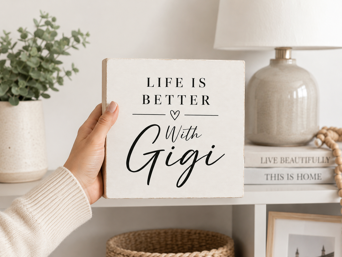 A hand in a cream knit sweater holds a square white decorative sign with the phrase “LIFE IS BETTER With Gigi” in black lettering. The sign is shown in a styled home interior setting with a shelf, lamp, books, and plant in the background.