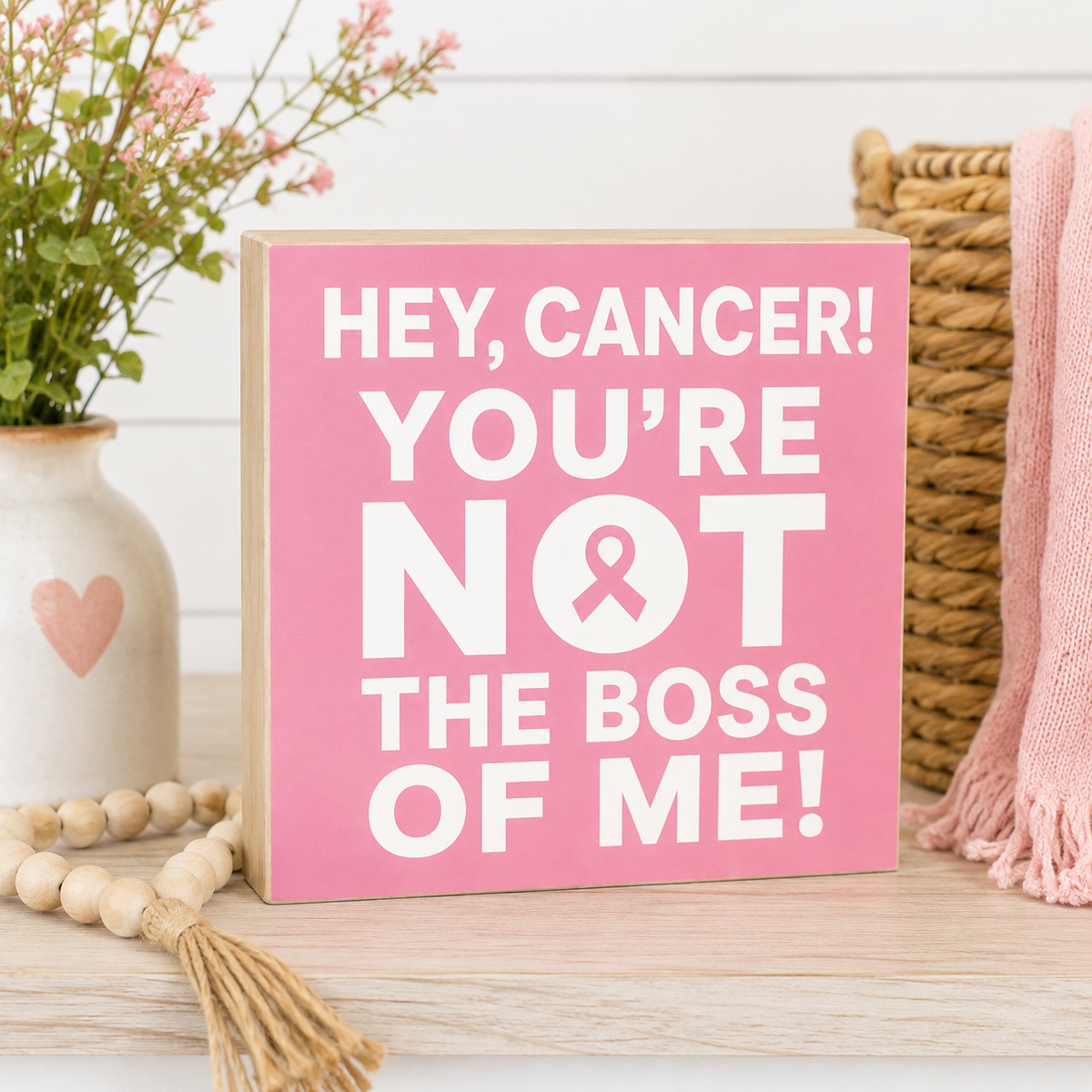 A square pink wooden sign with white text reading 'HEY, CANCER! YOU'RE NOT THE BOSS OF ME!' stands on a light wood surface. The sign is styled in a home setting with a white vase of flowers, wooden beads, a wicker basket, and a pink knit throw.