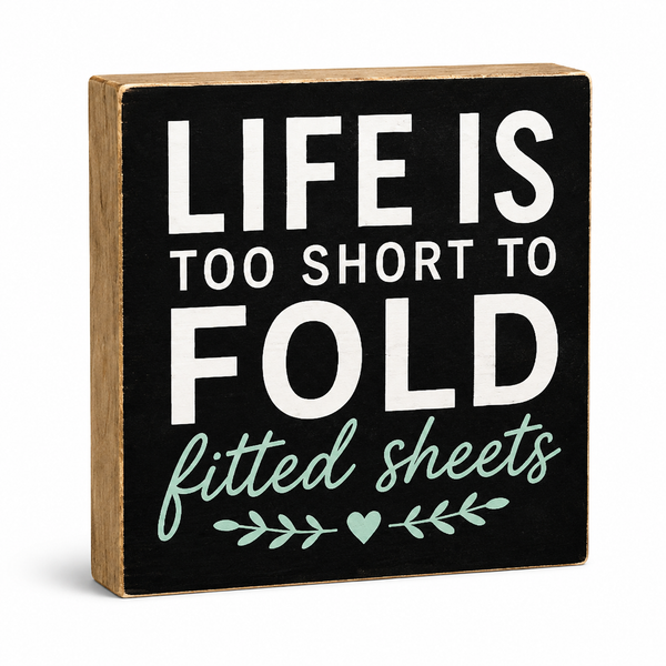 A square decorative wooden sign with a black front panel and natural wood sides. The sign reads “LIFE IS TOO SHORT TO FOLD” in large white letters, with “fitted sheets” in mint green script below, plus a small mint green leaf and heart motif.