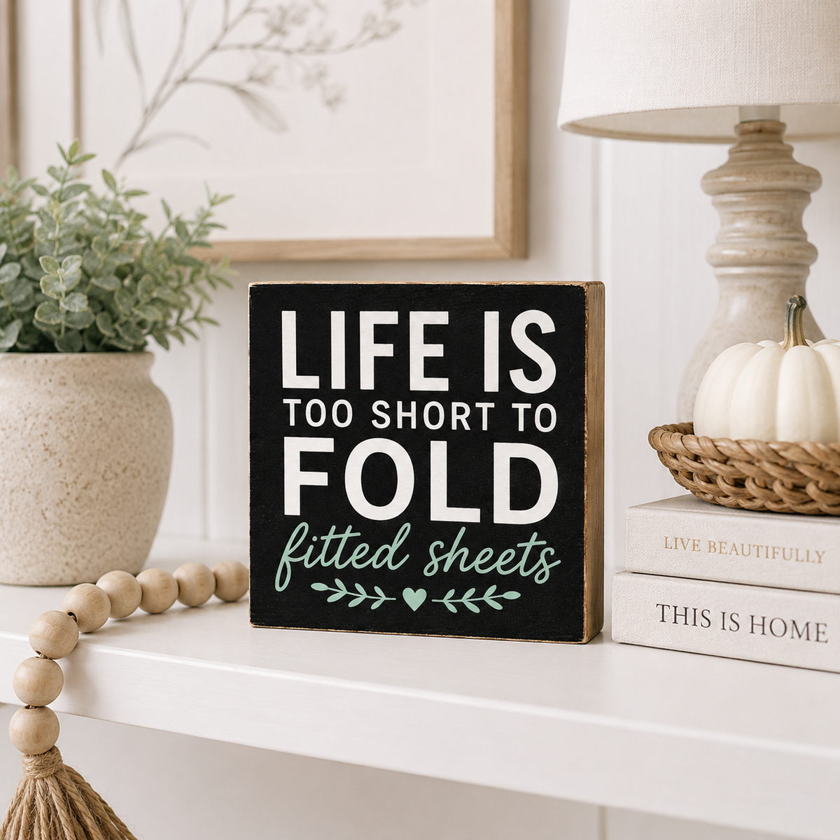 A rectangular black decorative sign with rustic wood edges and white text reading “LIFE IS TOO SHORT TO FOLD” with “fitted sheets” in teal script below, styled on a white shelf beside a lamp, books, a small pumpkin, and a plant.