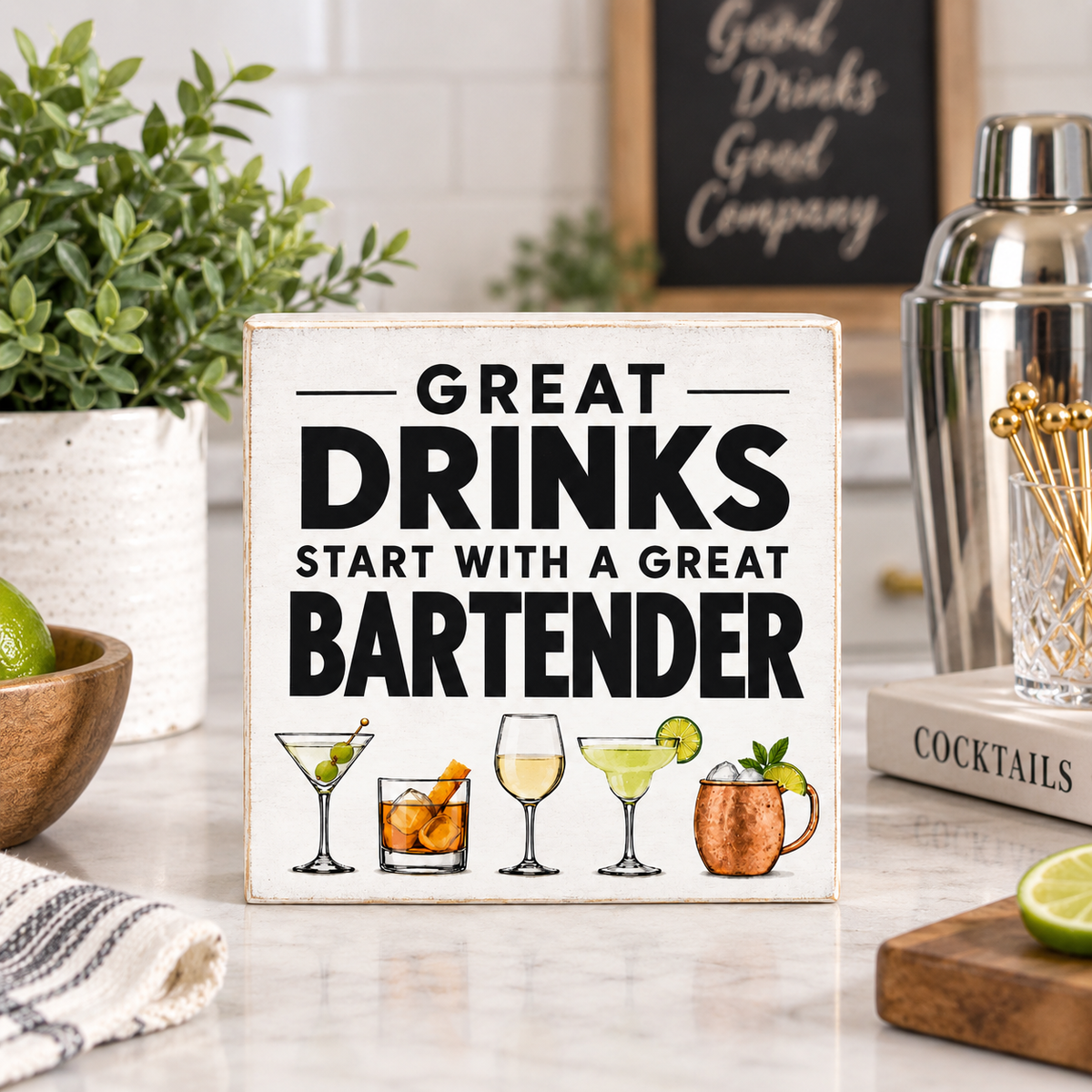A square decorative sign with the phrase “GREAT DRINKS START WITH A GREAT BARTENDER” in bold black text, surrounded by illustrated cocktail glasses and garnishes. The sign is displayed on a kitchen counter with a shaker, books, plants, and lime slices in a bright, modern kitchen setting.