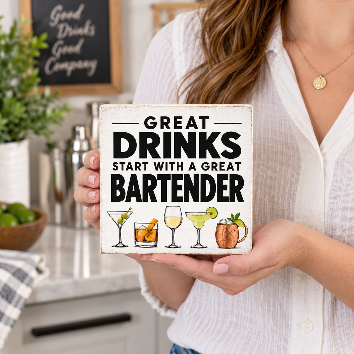 A woman in a white button-down shirt holds a square white decorative sign with the text 'GREAT DRINKS START WITH A GREAT BARTENDER' and small cocktail illustrations. The background shows a softly blurred kitchen or bar setting with a chalkboard sign, plant, and bar accessories.