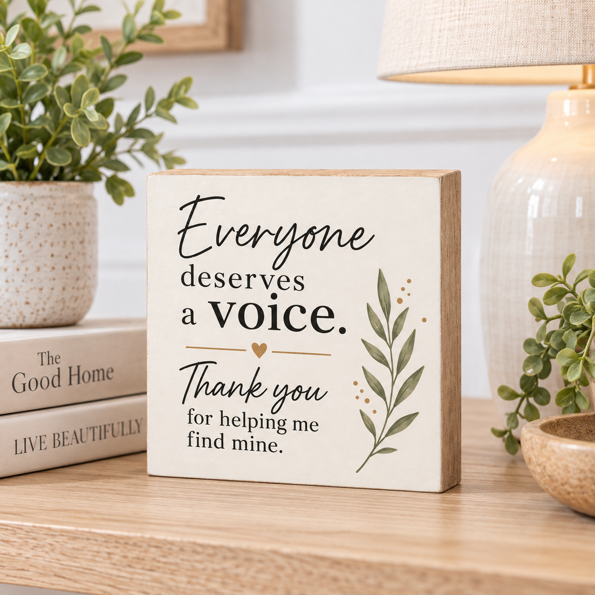 A square wooden sign with a cream face and natural wood edges, printed with the quote 'Everyone deserves a voice. Thank you for helping me find mine' in black script and serif text, with a small leaf illustration.