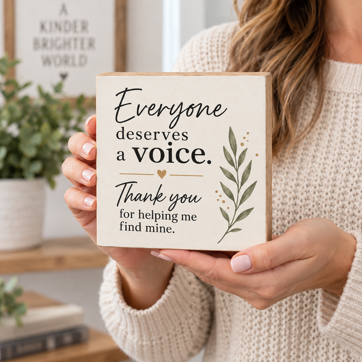 A woman in a cream knit sweater holds a square wooden sign that reads 'Everyone deserves a voice. Thank you for helping me find mine.