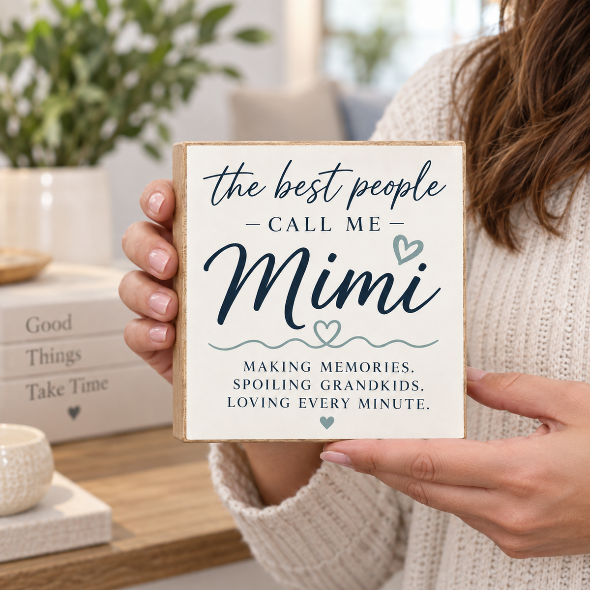A person in a cream knit sweater holds a square wooden sign that reads, 'the best people CALL ME Mimi MAKING MEMORIES. SPOILING GRANDKIDS, LOVING EVERY MINUTE.