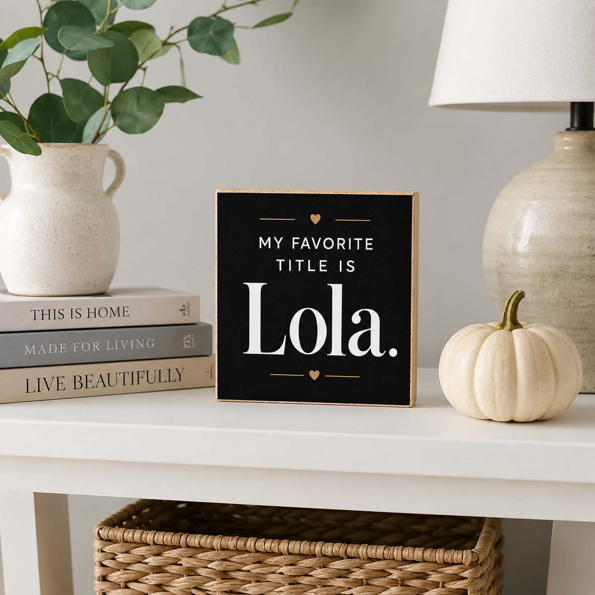A square black decorative sign with a light wood border and white text reading 'MY FAVORITE TITLE IS Lola.' sits on a white table in a styled home setting, surrounded by a small white pumpkin, books, a vase with greenery, and a lamp.