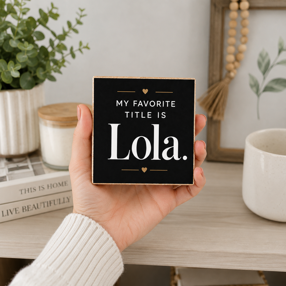 A hand in a white ribbed sweater holds a square black decorative sign with the text 'MY FAVORITE TITLE IS Lola.' in white lettering, set against a styled home interior background.