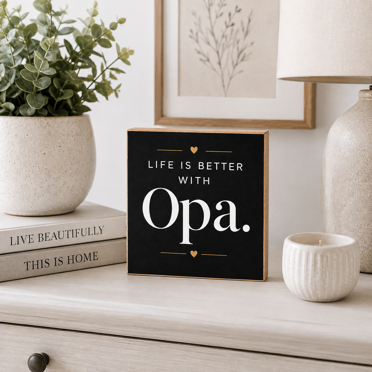 A square black decorative sign with light wood edges and white text reading 'LIFE IS BETTER WITH Opa.' styled on a light wood dresser beside a potted plant, books, a candle, and a lamp.