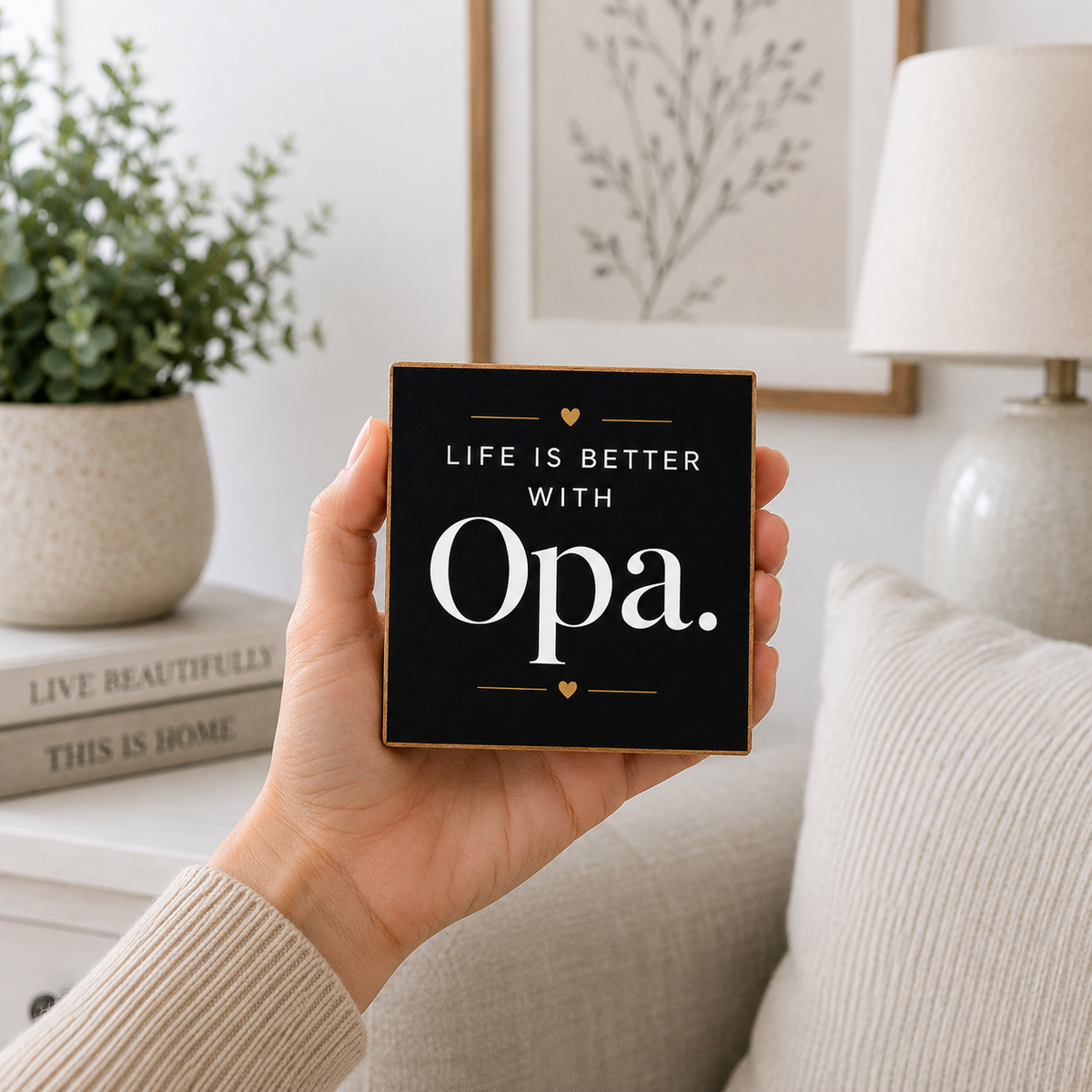 A hand holds a square black decorative sign with gold trim and white text that reads 'LIFE IS BETTER WITH Opa.' in a cozy living room setting with a sofa, lamp, and potted plant in the background.