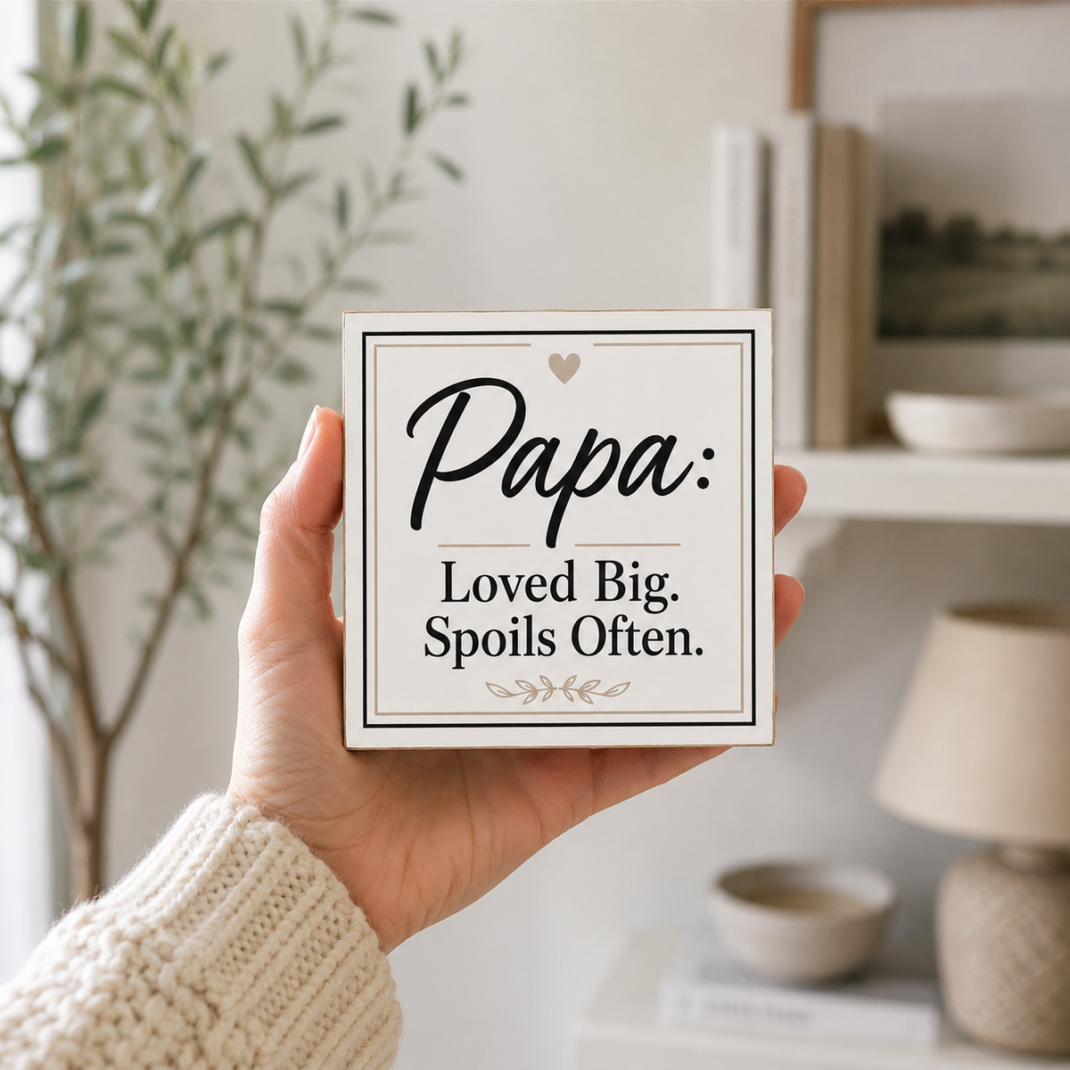 A hand wearing a cream knit sweater holds a square white sign with the text 'Papa: Loved Big. Spoils Often.