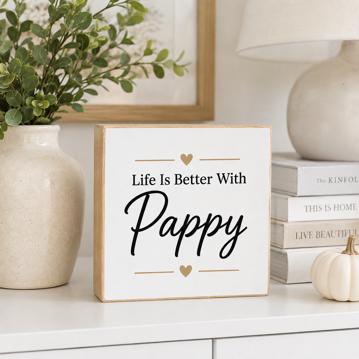 A square white decorative sign with a light wood border stands on a white shelf in a styled home setting. The sign reads “Life Is Better With Pappy” in black script and serif text, with small gold heart accents.