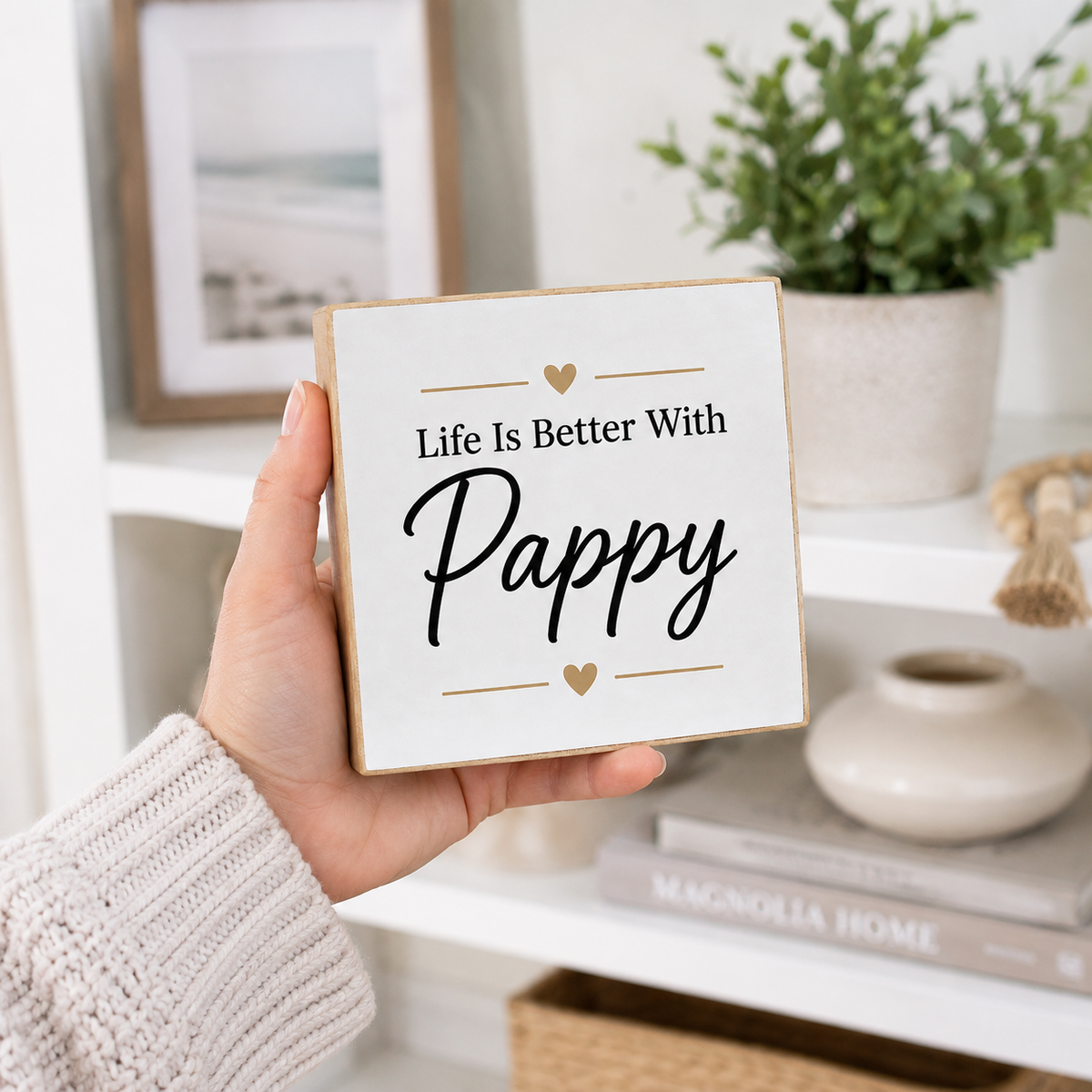A hand in a cream knit sweater holds a square white decorative sign with a natural wood border. The sign reads 'Life Is Better With Pappy' in black script and serif text, with small gold heart and line accents.