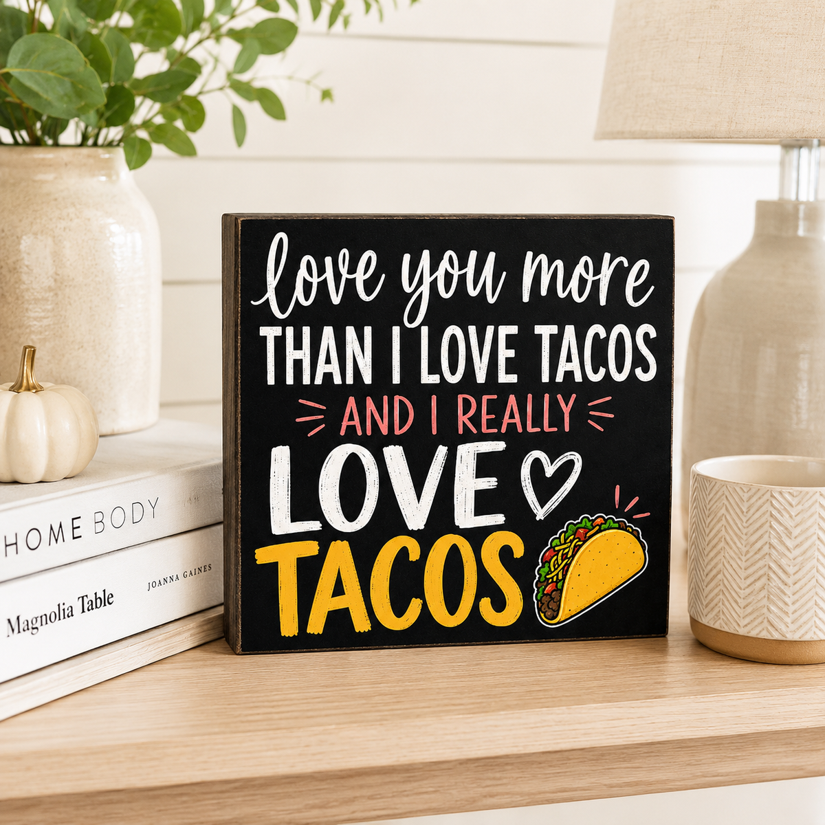 A square black decorative sign with white, pink, and yellow text reading 'love you more THAN I LOVE TACOS AND I REALLY LOVE TACOS' plus a taco illustration, displayed on a wooden shelf with books, a plant, and home decor in a styled interior setting.