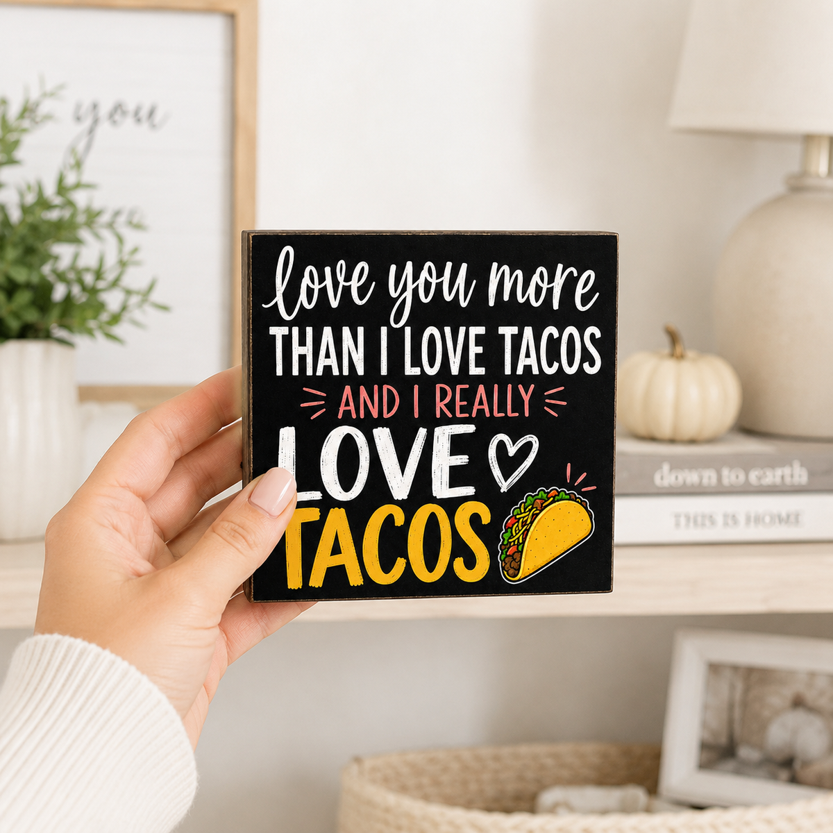 A hand holds a square black decorative sign with the phrase 'love you more THAN I LOVE TACOS AND I REALLY LOVE TACOS' in white, pink, and yellow lettering, alongside a taco illustration. The background shows a softly blurred home interior with a shelf, lamp, books, and small pumpkin.