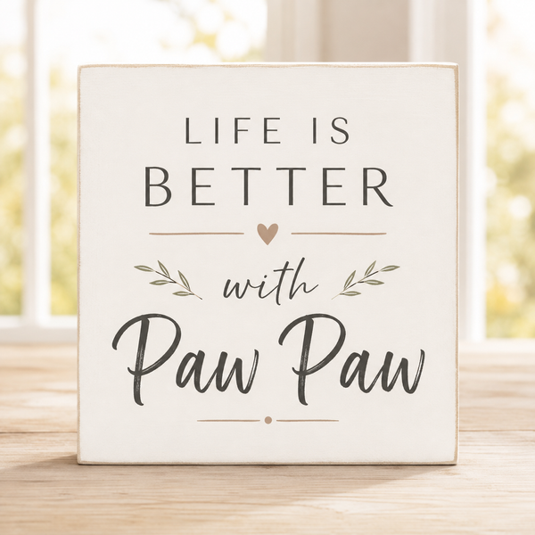A square cream-colored wooden sign with the text 'LIFE IS BETTER with Paw Paw' in dark gray lettering, featuring a small heart and leaf illustrations, displayed on a light wooden surface with a blurred window background.