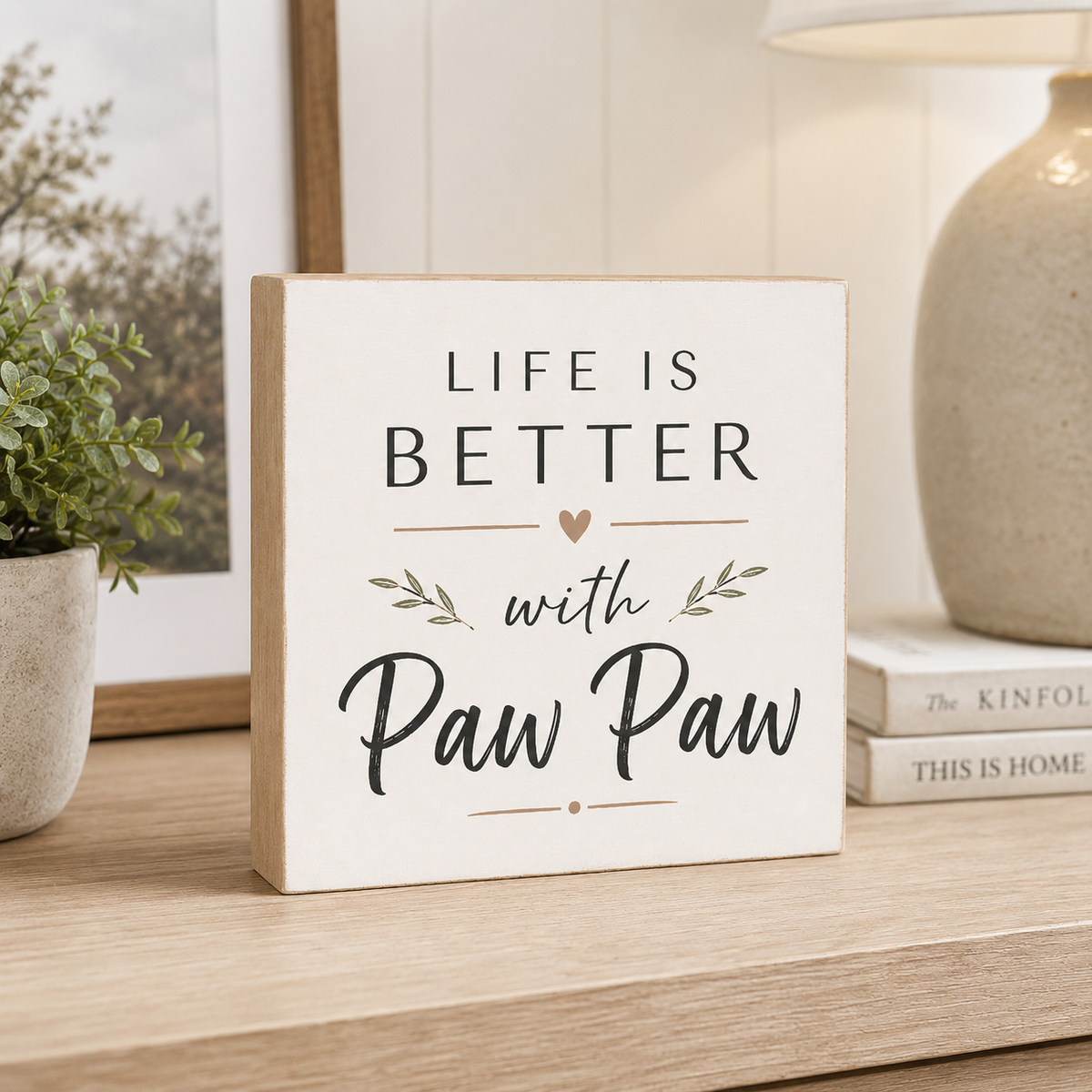 A square decorative wooden sign with the phrase 'LIFE IS BETTER with Paw Paw' in black lettering, styled on a light wood shelf next to a potted plant and a lamp.
