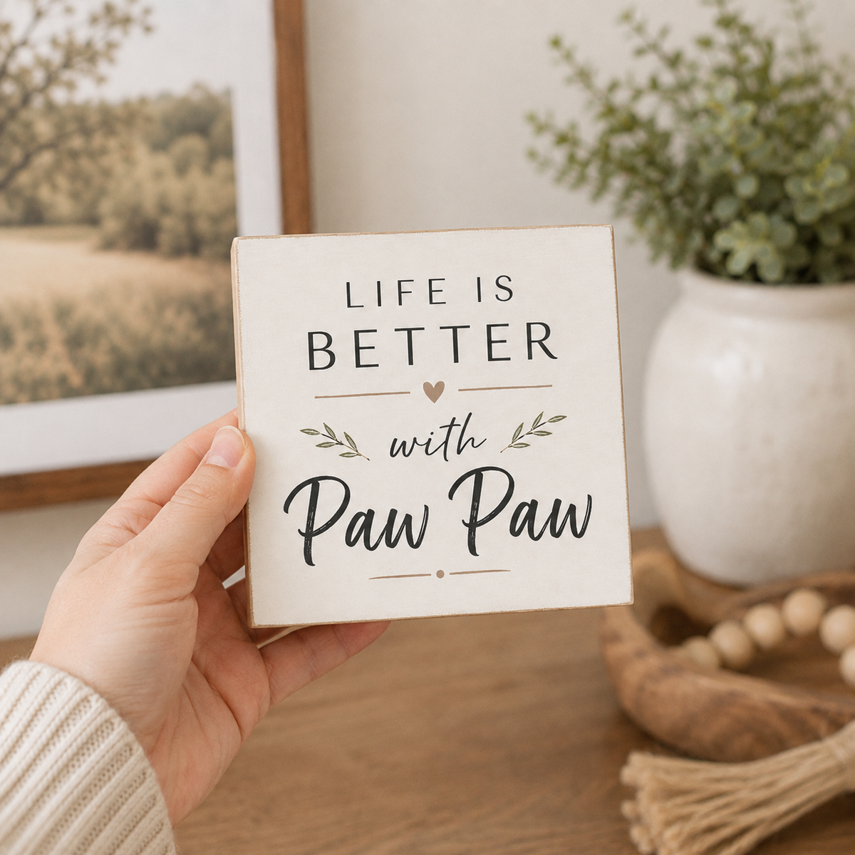 A hand holds a square decorative sign that reads 'LIFE IS BETTER with Paw Paw' in black lettering on a cream background. The sign has a light wood border and is photographed in a cozy indoor setting with a framed print, potted plant, and wooden decor in the background.