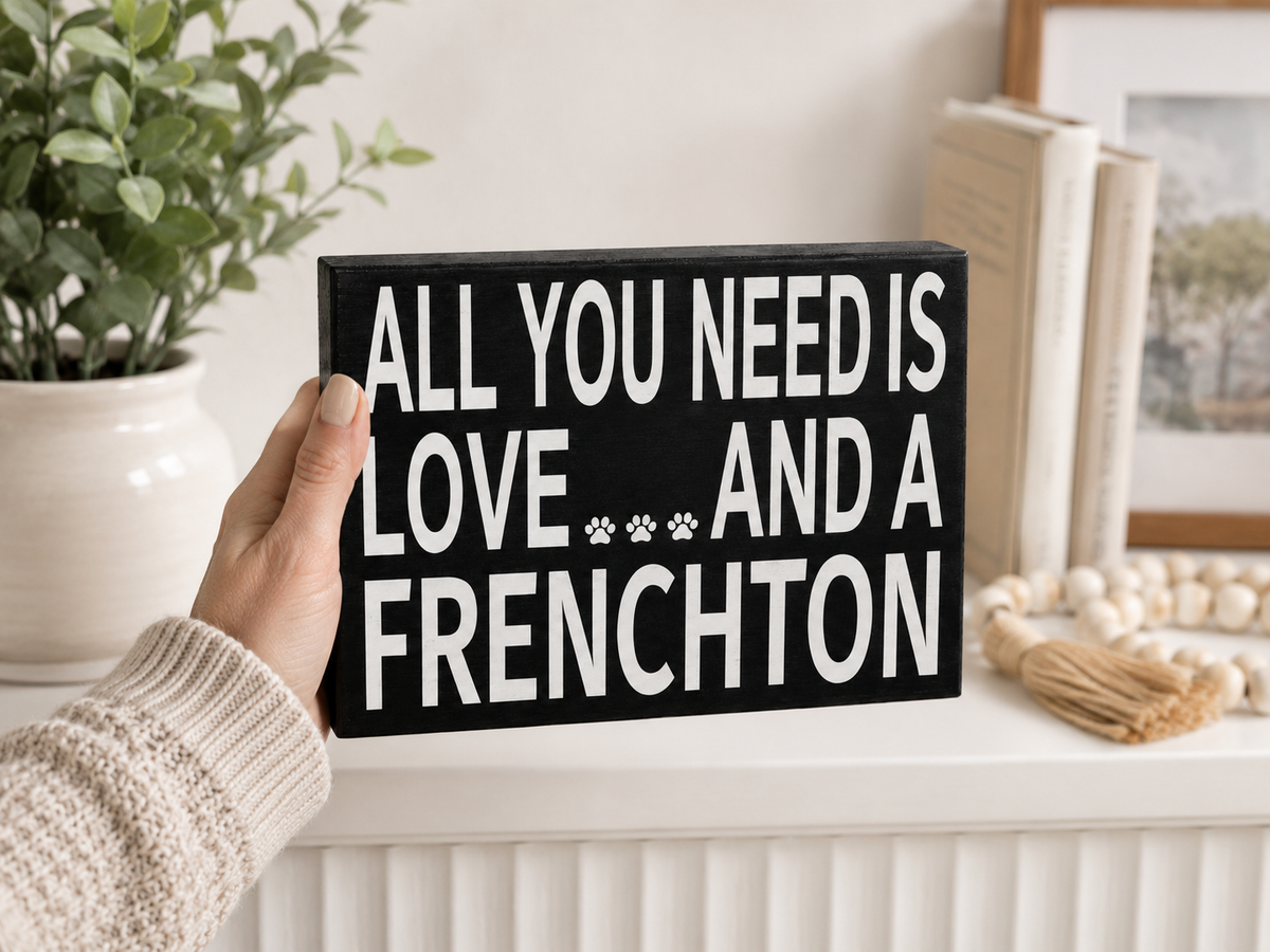 A hand in a cream knit sweater holds a black rectangular sign with bold white text reading 'ALL YOU NEED IS LOVE.