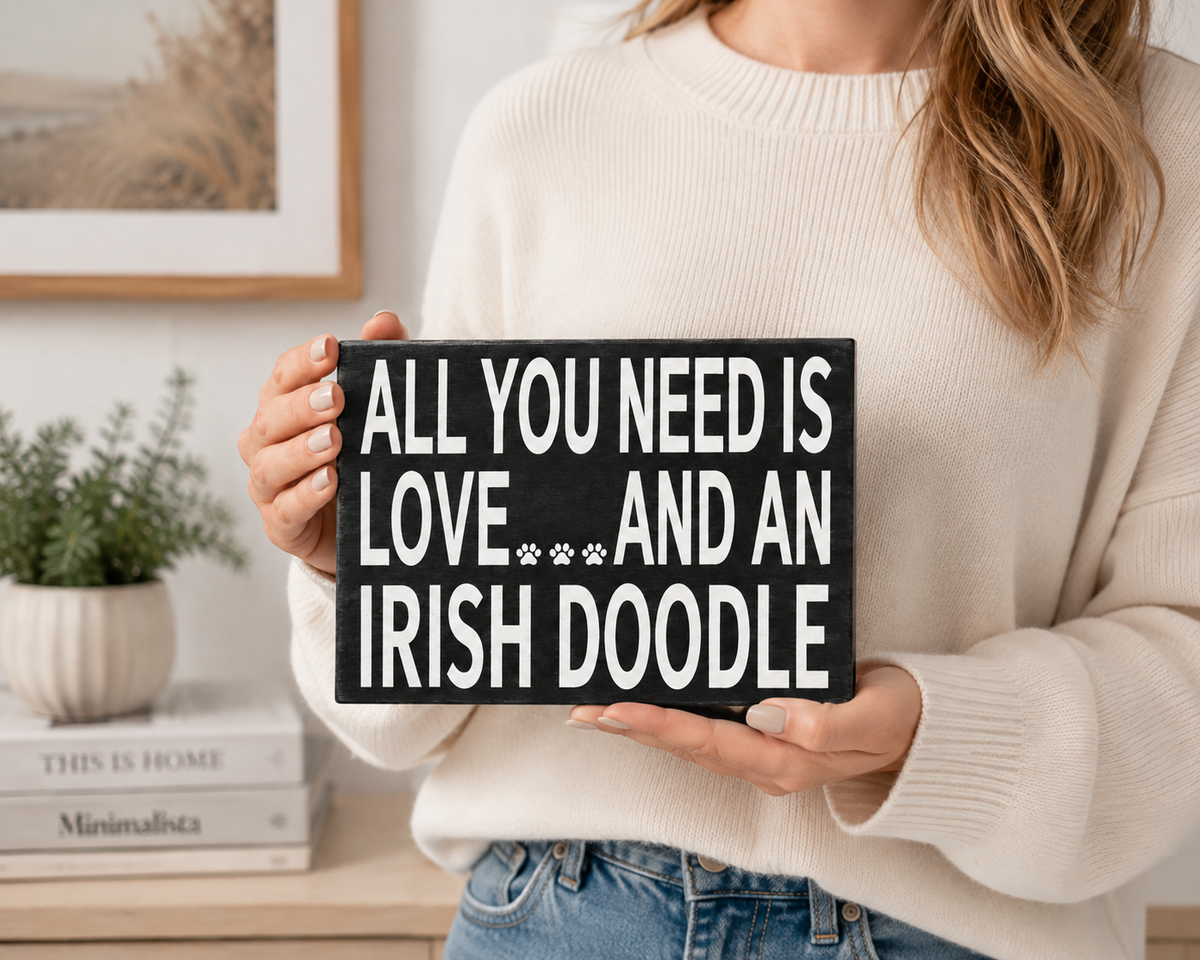 A person in a cream knit sweater holds a black rectangular sign with white text that reads, 'ALL YOU NEED IS LOVE.