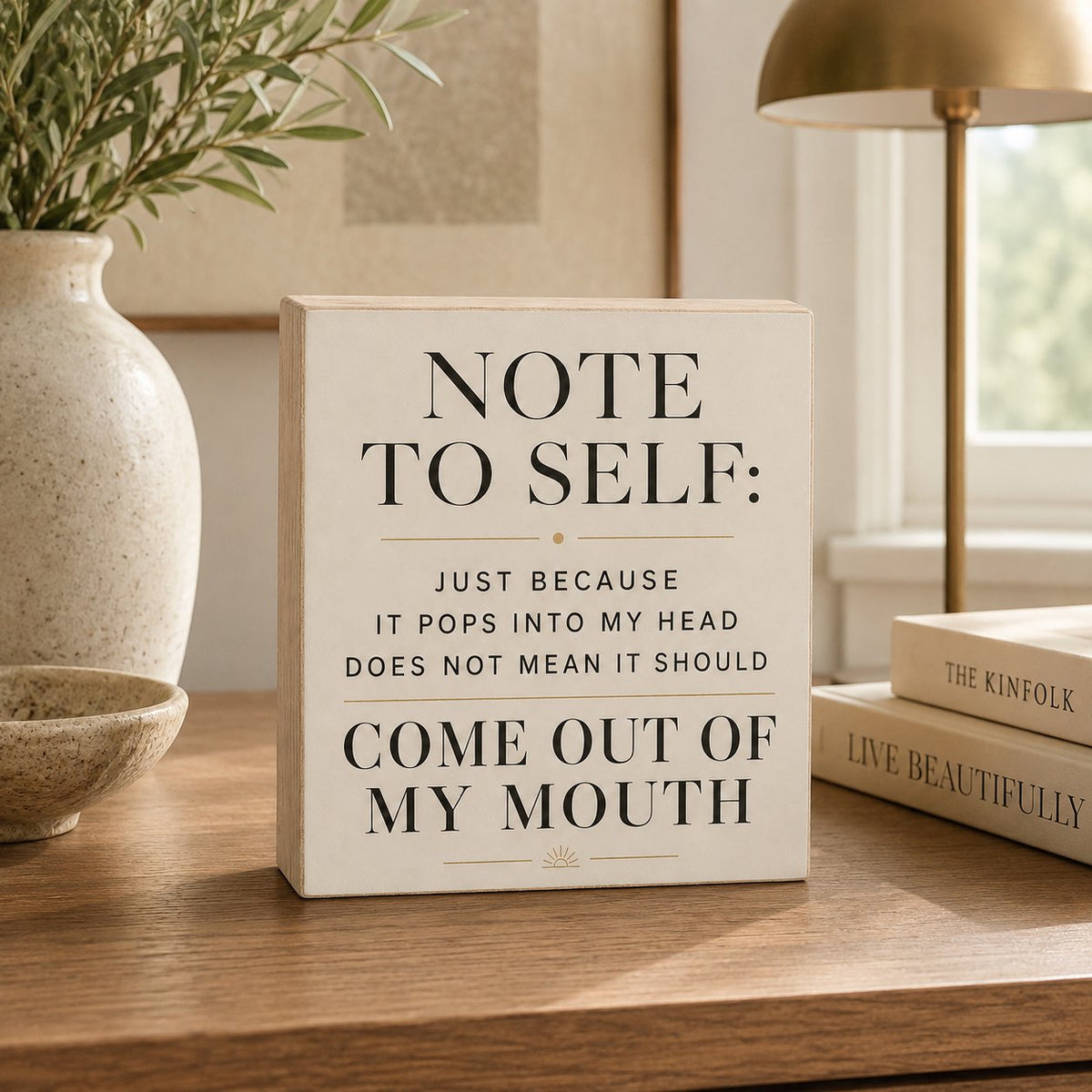 A square cream-colored decorative sign with black text reading 'NOTE TO SELF: JUST BECAUSE IT POPS INTO MY HEAD DOES NOT MEAN IT SHOULD COME OUT OF MY MOUTH' sits on a wooden table in a styled interior setting with a vase, books, and a lamp.