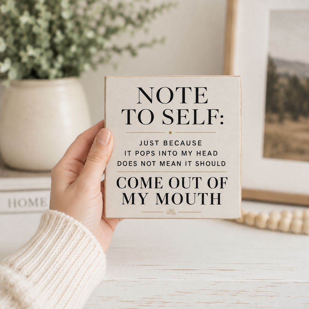 A hand in a cream knit sweater holds a square beige card with black text that reads, 'NOTE TO SELF: JUST BECAUSE IT POPS INTO MY HEAD DOES NOT MEAN IT SHOULD COME OUT OF MY MOUTH.' The background shows a softly blurred home interior with a potted plant, books, and framed art.