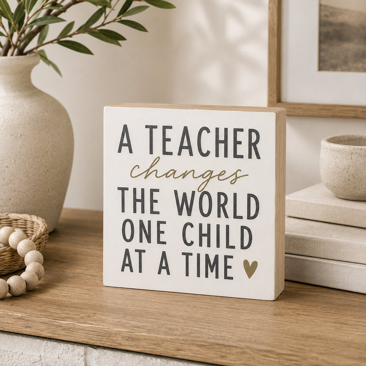 A square white wooden sign with a light natural wood edge, featuring the text 'A TEACHER changes THE WORLD ONE CHILD AT A TIME' in dark gray lettering with a small gold heart. The sign is styled on a wooden shelf next to a ceramic vase, books, and a bowl.
