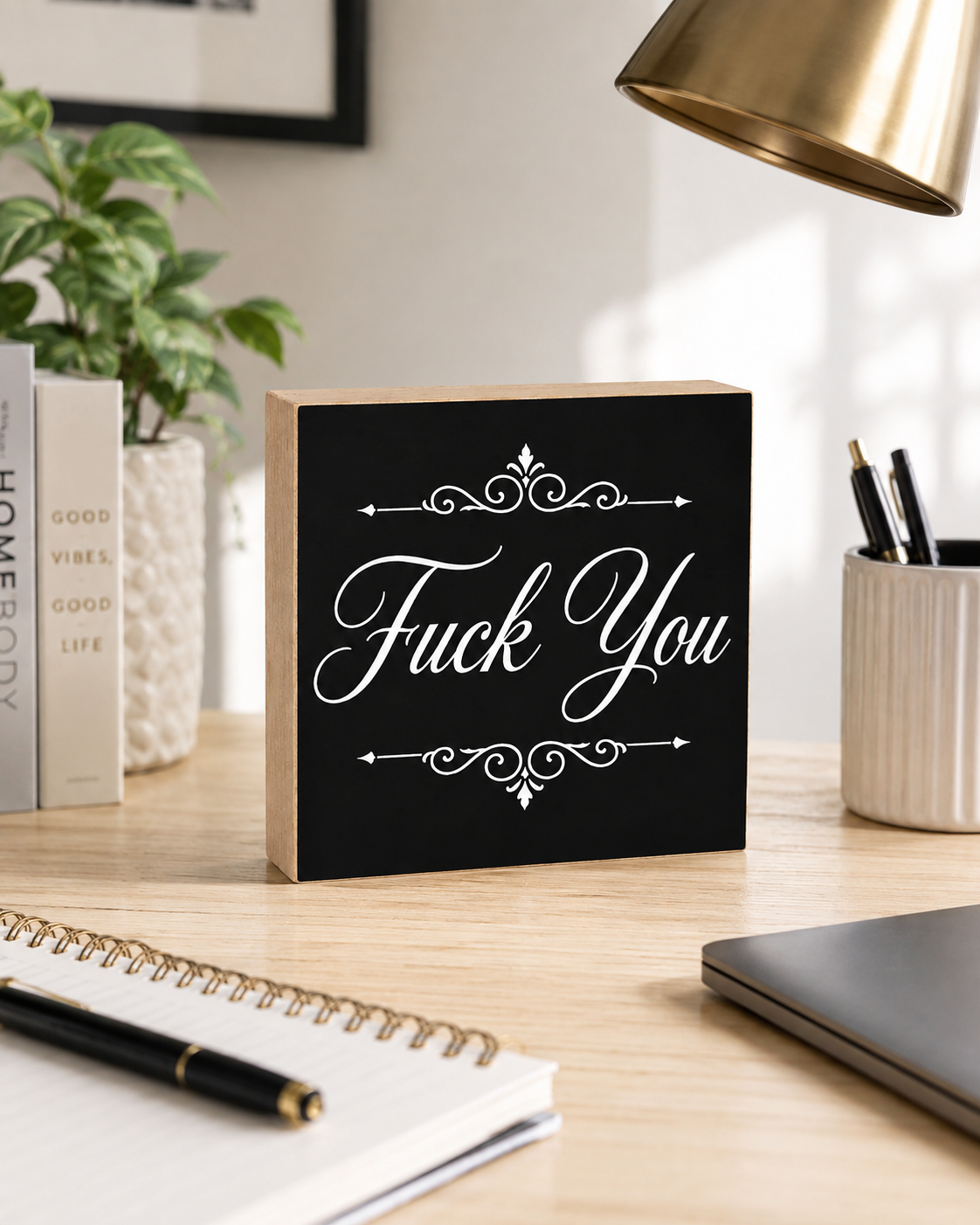 A square black decorative block sign with white script text reading 'Fuck You' and ornamental flourishes, displayed on a wooden desk in a styled home office setting with a plant, notebook, lamp, and laptop nearby.