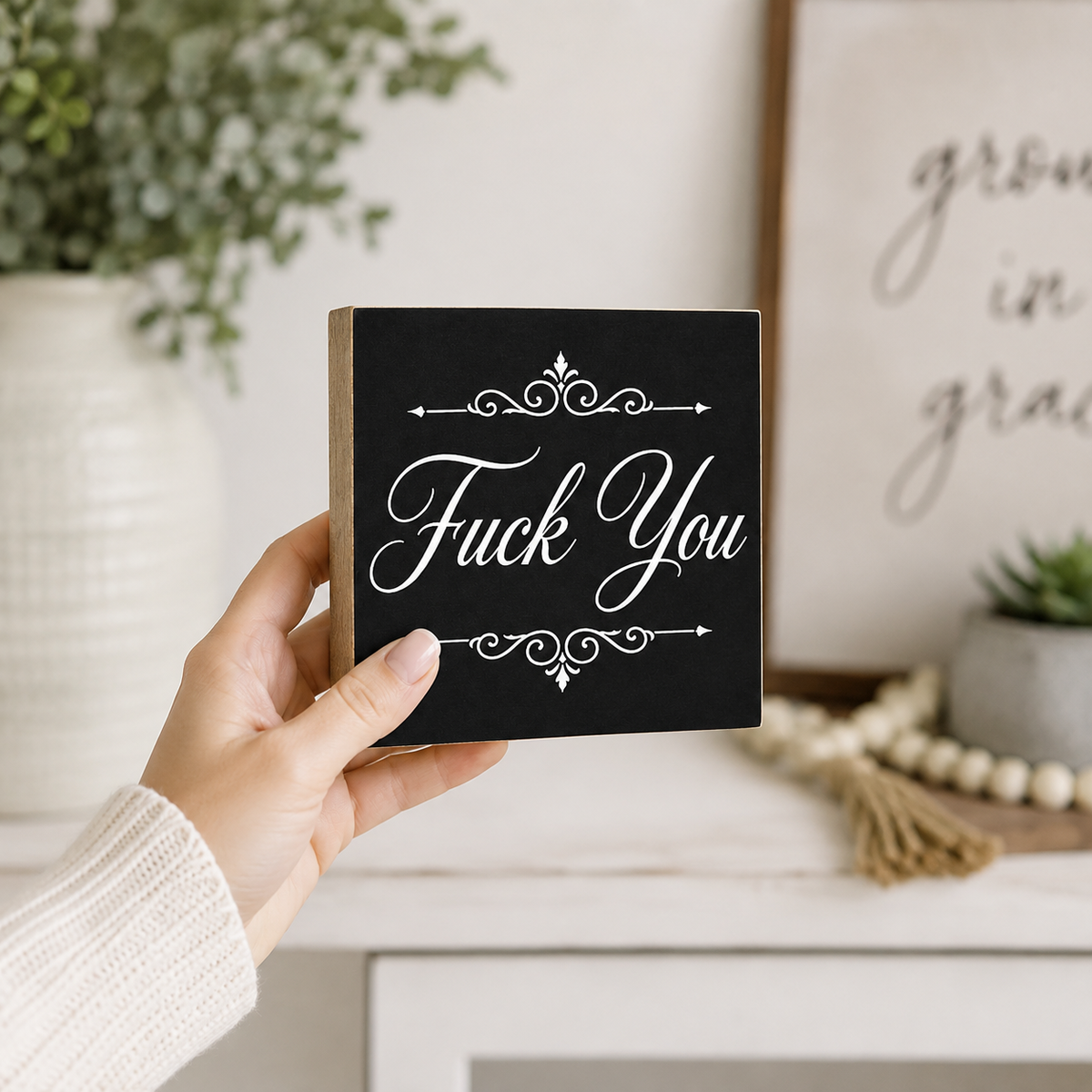 A hand holding a square black decorative block sign with white script text reading 'Fuck You' and ornamental flourishes, set against a softly blurred home interior background.