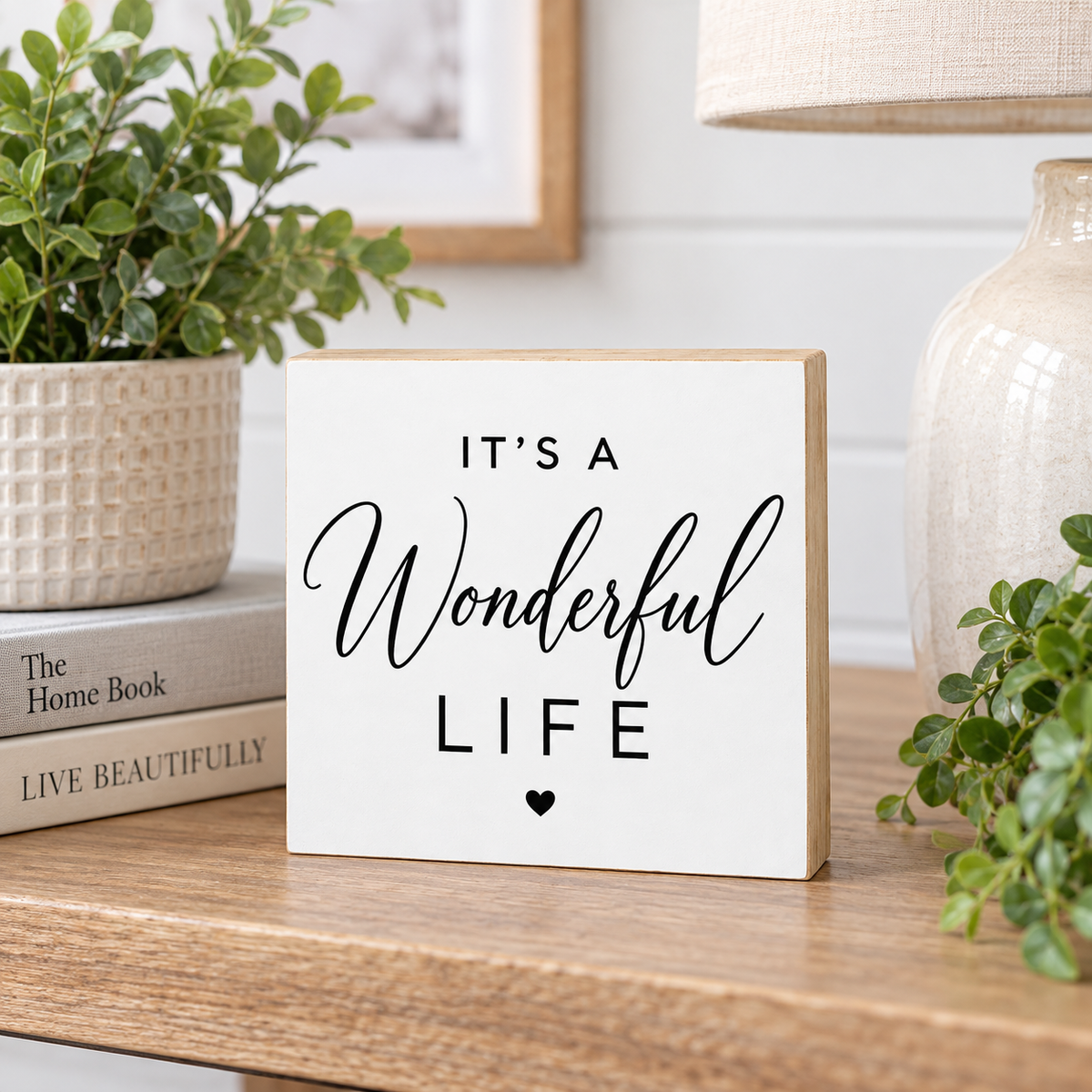 A square white decorative block sign with a light wood edge, printed with the phrase 'IT'S A Wonderful LIFE' in black lettering, displayed on a wooden shelf next to a potted plant and a lamp.