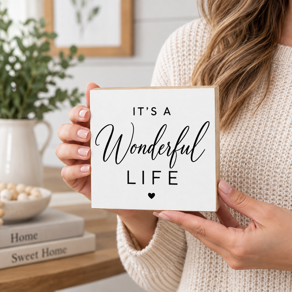 A woman in a cream knit sweater holds a square white sign with a light wood border. The sign reads 'IT'S A Wonderful LIFE' in black lettering with a small heart at the bottom.